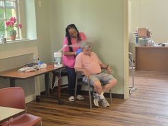 The image depicts another personal‑care session taking place in the same community‑center room. A seated participant receives a hair‑ or scalp‑related service from an individual in professional attire standing behind the chair. A nearby table displays treatment tools such as a towel, gloves, a spray bottle, and a device used in the session. The background includes a workspace with office furniture and organized supplies, showcasing a functional, multi‑purpose setting within the facility.