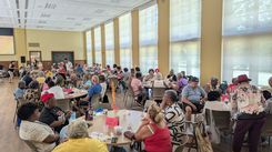 A wide-angle view of a large community event taking place in a spacious hall filled with round tables and many attendees. Each table is decorated with oversized paper flowers and colorful signage. People are seated, eating, chatting, or participating in group activities. Large windows line the wall, diffusing natural light across the room. A stage area is visible toward the left, where equipment and presenters appear to be set up. The atmosphere reflects a well-organized social gathering or celebratory luncheon.