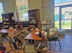 The image shows participants seated around tables inside a community room at the Hungry Hill Senior Center, eating food and engaging in conversation during the Spades Tournament event. The room features bookshelves, computer stations, informational brochures, and natural light from several large windows. Plates of food, drinks, and papers rest on the tables. The setting highlights senior social activities, community engagement, friendly competition, indoor recreation, and the welcoming environment provided by Department of Elder Affairs programming.
