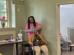 The image shows a community‑center wellness activity taking place in a softly lit room with pale green walls and large windows. A participant is seated in a chair while another individual, wearing professional attire and gloves, provides a hair‑ or scalp‑related service. A small table beside them holds care supplies, including a towel, a spray bottle, gloves, and a compact device that appears to be part of the treatment setup. A potted plant decorates the windowsill, and the setting overall resembles an organized, calm environment designed for health or personal‑care sessions.
