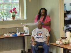This image captures another personal‑care session in the same community room. A participant sits in a chair while an individual standing behind provides a scalp or hair‑care service. A misting or steaming device is actively running beside them, positioned on a table that holds various small supplies such as gloves, cloths, and bottled items. A window in the background features potted flowers, adding color to the calm, organized setting.