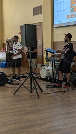 The image captures a live musical performance inside a community event hall. A vocalist stands at a microphone while nearby a musician plays an electronic keyboard. Behind them, additional performers are positioned near a drum set and sound equipment. The space includes wooden flooring, large speakers, and a projection screen displaying digital content. The scene reflects a structured indoor entertainment setup as part of a larger community event. Malado