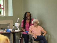 The third image shows a seated participant receiving a care treatment from an individual standing behind in a professional setting. The treatment appears to involve scalp or hair care, supported by a steaming device positioned on a nearby table. The surrounding area features neatly arranged supplies, a water bottle, and a window with decorative plants. The room’s soft‑colored walls and wooden floors create a warm, community‑center environment.