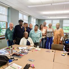 A group gathered around a table in the same activity room, standing together for a group photo. The table displays craft supplies, magazines, scissors, and personal items. The room has multiple large windows and overhead lighting.