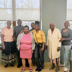 A group of individuals standing together in a room with large windows in the background. The group is arranged in a line, shoulder‑to‑shoulder, and appears to be participating in an indoor community gathering or class. The room has pale green walls, light flooring, and natural light coming through the windows. Some individuals are wearing casual clothing, while others are dressed more formally. One mobility walker is visible to the right.