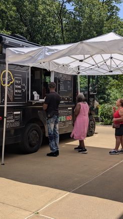 The image shows an outdoor food‑truck setup under a white canopy tent. Several individuals wait in line at a black ice‑cream or dessert truck featuring flavor menus, chalkboard signage, and promotional graphics. The truck is parked on a paved walkway bordered by tall green trees, giving the scene a relaxed, summer event atmosphere. A small counter extends from the service window where orders are being taken, and the surrounding area appears clean, organized, and event‑ready.