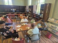 The image shows participants seated around tables inside a community room at the Hungry Hill Senior Center, eating food and engaging in conversation during the Spades Tournament event. The room features bookshelves, computer stations, informational brochures, and natural light from several large windows. Plates of food, drinks, and papers rest on the tables. The setting highlights senior social activities, community engagement, friendly competition, indoor recreation, and the welcoming environment provided by Department of Elder Affairs programming.