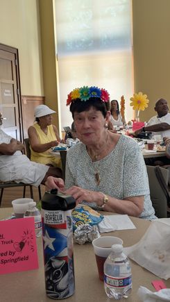 The image shows a participant seated at a decorated table inside a large event space. The individual is wearing a colorful floral headband and is engaged in an activity at the table. Surrounding the person are event materials including snack bags, water bottles, cups, and a patterned water container. A bright paper flower centerpiece and pink event signage are positioned nearby. Other attendees can be seen seated at adjacent tables, contributing to a lively community gathering environment.