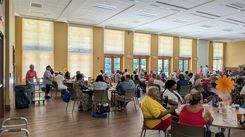 A view of a crowded community hall where numerous attendees are seated around tables enjoying a meal. The tables are decorated with large paper flowers and event markers. The room has tall windows, wooden flooring, and warm-colored walls that create an inviting ambiance. Individuals are engaged in conversation, dining, or participating in the event’s activities. Staff can be seen moving through the space, and the overall setting suggests a coordinated and lively community event.
