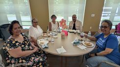 A group of individuals sits around a round table in a bright community‑center room enjoying a meal. The table is set with plates of food, bottled water, small snack bags, and cups of beverages. A large decorative paper flower stands in the center beside a table number card and a pink event label. The background features large windows with frosted coverings, allowing natural light to fill the room. The atmosphere appears organized and social, suggesting a community luncheon or celebration.