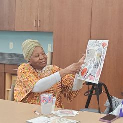 An individual seated at a table in a craft or activity room holding up a collage board featuring cut‑out pictures, text, and decorative elements. The table has craft supplies such as markers, glue sticks, cups, and paper.