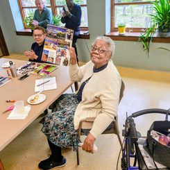 An individual seated at a table holding up a completed collage featuring travel‑related images and text. On the table are snacks, a drink, craft tools, and a mobility walker positioned nearby.