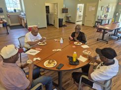 The image shows participants seated around tables inside a community room at the Hungry Hill Senior Center, eating food and engaging in conversation during the Spades Tournament event. The room features bookshelves, computer stations, informational brochures, and natural light from several large windows. Plates of food, drinks, and papers rest on the tables. The setting highlights senior social activities, community engagement, friendly competition, indoor recreation, and the welcoming environment provided by Department of Elder Affairs programming.
