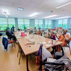 A large room with several people seated around long tables, each working on collage projects. The tables are covered with magazines, papers, scissors, glue, and finished art boards. Large windows line the wall, allowing bright natural light into the space.