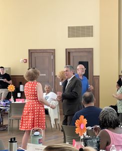 The image highlights a closer view of the same community event. Several attendees are standing and talking near the center of the room, while others remain seated at decorated round tables. Large paper flowers serve as table centerpieces, and food items are visible on the tables. The background shows neutral-colored walls, wooden doors, and additional attendees moving through the room. The setting maintains a structured, community‑focused environment with a festive, social ambiance.