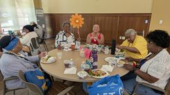 A group of individuals sits around a circular table during a community luncheon event. The table is covered with plates of food, bottled water, wrapped items, drink cups, and assorted snacks. At the center of the table is a tall decorative flower and table number sign, surrounded by additional event décor including a pink label card. The room features warm-toned walls and large windows that allow natural light to brighten the space. Other attendees are seated at nearby tables in the background, enjoying their meals.